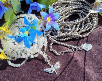 Op de afbeelding: Twee sets kralen sieraden met zilveren bedels. De ene set is wit en de andere is zilver. De sieraden liggen op een donkerbruin oppervlak met blauwe bloemen.