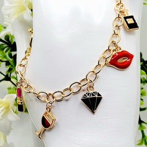 Può includere: Un braccialetto con ciondoli dorati con una varietà di ciondoli, tra cui un rossetto, un bicchiere di vino, un diamante, un paio di labbra e un flacone di profumo.