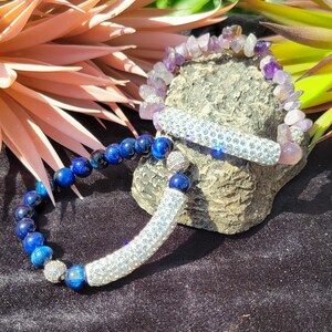 Pode incluir: Duas pulseiras com detalhes prateados e de cristal. Uma pulseira é feita de contas azuis de olho de tigre e a outra é feita de contas de ametista roxa.