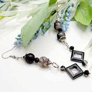 May include: A pair of silver earrings with black and gray agate beads and silver accents. The earrings have a diamond-shaped pendant with a clear center.