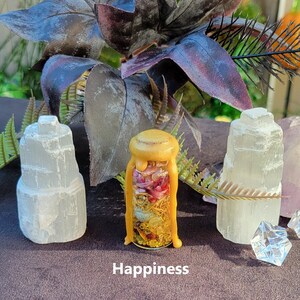 May include: A glass bottle with a yellow wax seal, filled with dried flowers and herbs, sits between two white selenite crystal towers. The word "Happiness" is printed at the bottom. The background includes dark purple leaves and greenery.