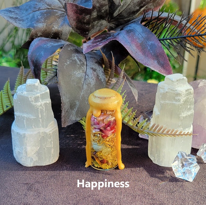 May include: A glass bottle with a yellow wax seal, filled with dried flowers and herbs, sits between two white selenite crystal towers. The word "Happiness" is printed at the bottom. The background includes dark purple leaves and greenery.