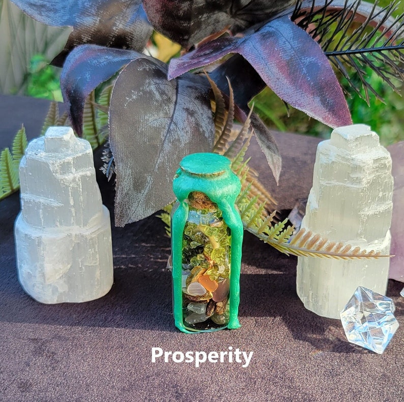 May include: A small glass bottle filled with colorful stones and sealed with green wax. The word "Prosperity" is printed at the bottom. Two white selenite towers and a clear crystal are on either side of the bottle, with dark purple leaves in the background.