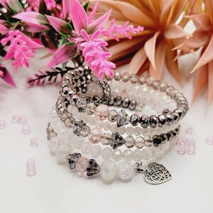 Puede incluir: Un brazalete de múltiples hilos con cuentas plateadas, blancas y rosadas. El brazalete tiene un dije de corazón plateado.