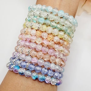 May include: A stack of colorful bracelets made of small, clear, crackled glass beads. The bracelets are layered on a person's wrist, creating a rainbow effect.