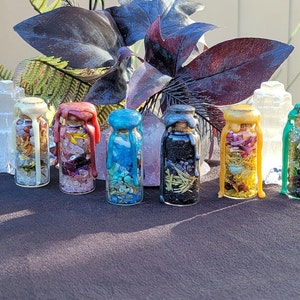 May include: A collection of seven small glass bottles filled with various dried herbs, crystals, and other decorative elements. Each bottle is sealed with a cork and colorful wax. The bottles are arranged on a dark surface with foliage in the background.