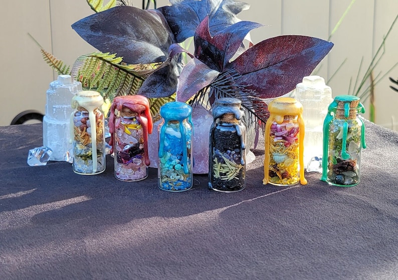 May include: A collection of seven small glass bottles filled with various dried herbs, crystals, and other decorative elements. Each bottle is sealed with a cork and colorful wax. The bottles are arranged on a dark surface with foliage in the background.
