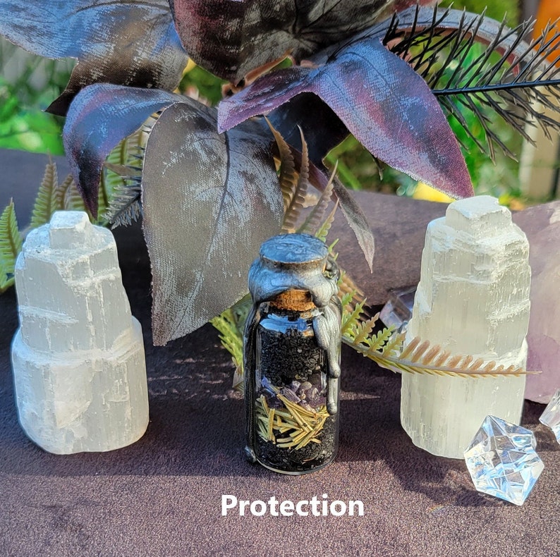 May include: A small glass bottle with a silver-colored lid and a cork stopper, filled with black and purple herbs and spices. The word "Protection" is printed at the bottom. Two white selenite towers flank the bottle, with greenery in the background.