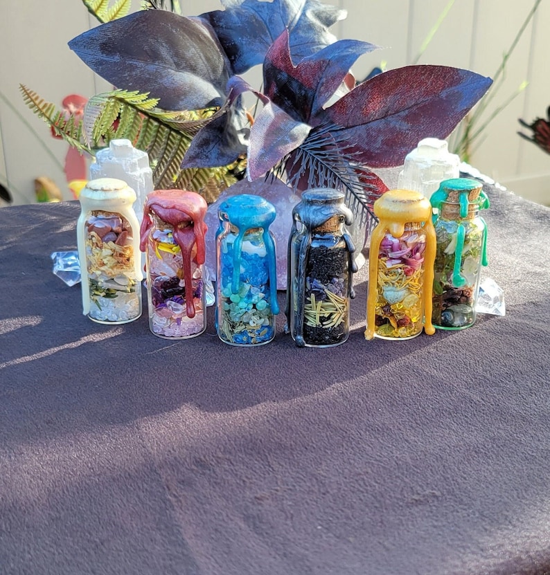 May include: Six small glass bottles filled with colorful herbs, crystals, and oils. Each bottle has a cork stopper sealed with wax in various colors, including white, red, blue, silver, yellow, and green. The bottles are arranged on a dark surface.