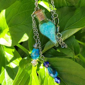 Op de afbeelding: Een zilveren ketting met een kleine glazen fles hanger gevuld met blauwe kristallen. De fles heeft een kurk. Extra blauwe kristallen en kralen accenten hangen aan de ketting, tegen een achtergrond van groene bladeren.