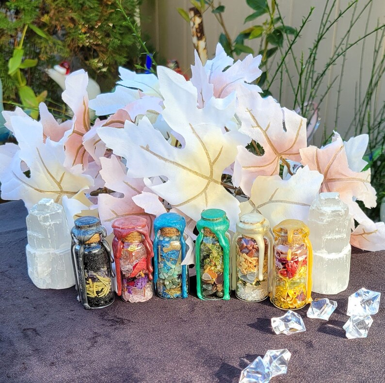 May include: A collection of small glass bottles filled with colorful crystals and dried flowers. Each bottle is sealed with a cork and wax in various colors, including black, red, blue, and green. The bottles are arranged in a row, with selenite crystals on either side.