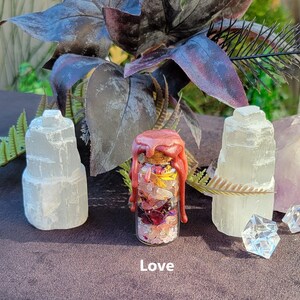 May include: A small glass bottle filled with colorful crystals and dried flowers, sealed with a cork and pink wax. The word "Love" is printed at the bottom. Two white selenite towers flank the bottle, with decorative foliage in the background.