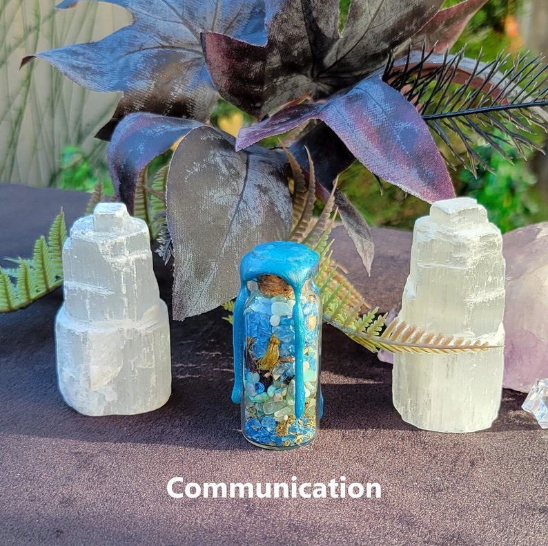 May include: A small glass bottle filled with blue and green stones, sealed with a blue wax top, flanked by two white selenite towers. The word "Communication" is printed at the bottom of the image. The background features dark purple leaves and greenery.
