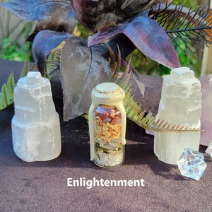 May include: A small glass jar filled with colorful stones and dried herbs, sealed with a creamy wax. Two white selenite towers flank the jar, with the word "Enlightenment" at the bottom. The background features dark purple foliage.