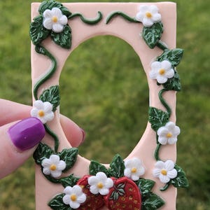 Könnte beinhalten: Ein rechteckiger, hellrosa Rahmen mit ovaler Öffnung, verziert mit weißen Blüten, grünen Blättern und Ranken. Zwei rote Erdbeeren mit weißen Samen befinden sich am unteren Rand. Der Rahmen wird von einer Hand mit einem lila lackierten Nagel gehalten.