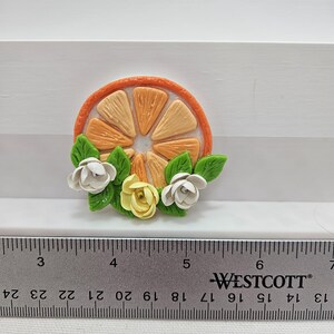 Pode incluir: Uma fatia de laranja artesanal com flores brancas e amarelas e folhas verdes. A fatia de laranja &eacute; rodeada por uma casca de laranja. Uma r&eacute;gua com medidas em cent&iacute;metros est&aacute; em primeiro plano.