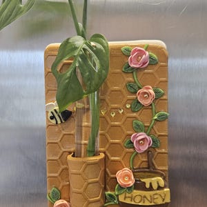 May include: A decorative wall planter with a honeycomb design, a bee, and a honey pot. A clear glass tube holds a green plant with a large leaf. Pink and peach roses and green leaves decorate the planter.