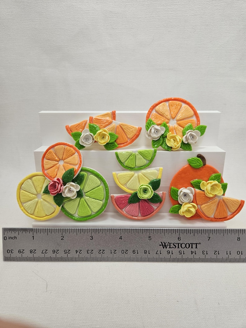 Pode incluir: Uma cole&ccedil;&atilde;o de ornamentos artesanais de fatias de frutas, incluindo laranja, lim&atilde;o, lima e toranja, cada um adornado com pequenos detalhes florais. A r&eacute;gua mostra um comprimento de cerca de 20 cm.