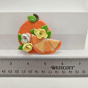 Pode incluir: Broche artesanal em forma de fatia de laranja, na cor laranja, com detalhes florais. O broche apresenta uma fatia de laranja, folhas verdes e flores brancas e amarelas. O broche mede aproximadamente 5 cm de comprimento.