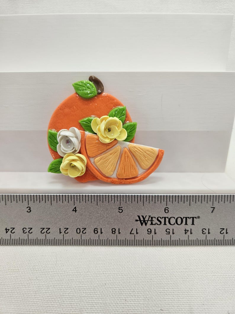 Pode incluir: Broche artesanal em forma de fatia de laranja, na cor laranja, com detalhes florais. O broche apresenta uma fatia de laranja, folhas verdes e flores brancas e amarelas. O broche mede aproximadamente 5 cm de comprimento.
