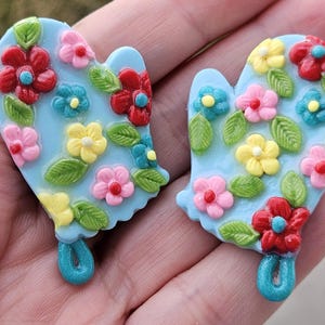 May include: Two light blue oven mitt-shaped decorations. Each mitt is decorated with colorful flowers in red, pink, yellow, and blue, along with green leaves. The mitts have a loop at the bottom.