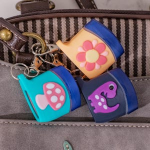 Puede incluir: Tres llaveros coloridos con diseños únicos: una flor, una seta y un dinosaurio. Cada llavero tiene una parte superior azul y un color base diferente. Los llaveros están sujetos a un cierre metálico y descansan sobre un bolso gris.