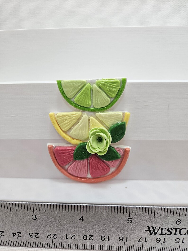 Pode incluir: Tr&ecirc;s decora&ccedil;&otilde;es coloridas de fatias de frutas: uma lima verde, um lim&atilde;o amarelo e uma toranja rosa. Uma pequena flor verde e folhas adornam as fatias de lim&atilde;o e toranja. As fatias de fruta s&atilde;o dispostas sobre uma superf&iacute;cie branca.