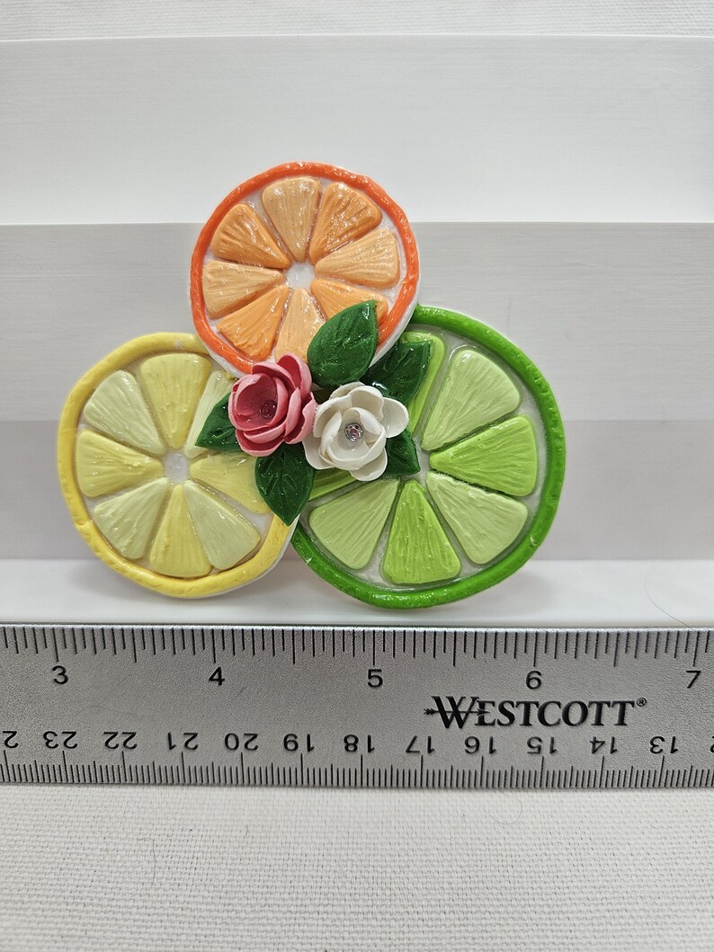 Pode incluir: Um broche colorido com tema c&iacute;trico apresentando fatias de laranja, lim&atilde;o e lima. O broche &eacute; adornado com pequenas flores de tecido e folhas verdes. Uma r&eacute;gua est&aacute; em primeiro plano, com a marca "WESTCOTT" vis&iacute;vel.