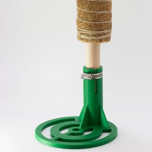 May include: A green plastic plant stand with a round base and a vertical support. The support has a metal clamp to hold a plant stake. The stake is wrapped in brown coconut fibre.