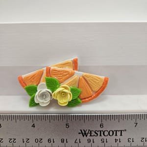 Pode incluir: Uma fatia decorativa de laranja com flores brancas e amarelas e folhas verdes. A fatia de laranja tem aproximadamente 5 cm de comprimento. Uma r&eacute;gua Westcott est&aacute; em primeiro plano.