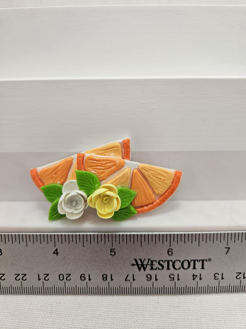 Pode incluir: Uma fatia decorativa de laranja com flores brancas e amarelas e folhas verdes. A fatia de laranja tem aproximadamente 5 cm de comprimento. Uma r&eacute;gua Westcott est&aacute; em primeiro plano.