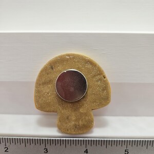 Pode incluir: Um &iacute;man em forma de cogumelo com um disco prateado met&aacute;lico na parte de tr&aacute;s. O &iacute;man &eacute; castanho com uma textura brilhante e um contorno amarelo. Uma r&eacute;gua est&aacute; na parte inferior, mostrando medidas em cent&iacute;metros.