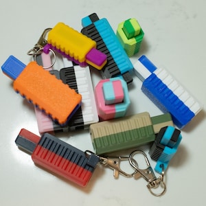 May include: A selection of brightly coloured, geometric keyrings in diverse shapes and sizes. The keyrings display a range of colours, including orange, yellow, blue, and green. Some are rectangular, while others are cube-shaped. Each keyring has a metal clasp.