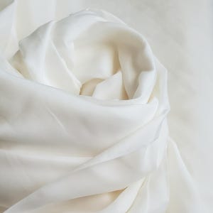 Può includere: Primo piano di un tessuto bianco con morbide pieghe e una texture setosa. Il tessuto è di un bianco crema, con riflessi e ombre che creano profondità. Il materiale appare leggero e fluido.