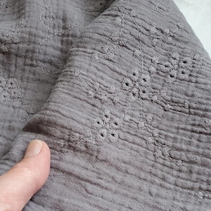May include: A piece of grey crinkled fabric featuring a delicate tonal embroidered floral pattern. The textile has a soft, textured appearance with a raw, unfinished edge visible along the bottom, showcasing the intricate stitching details.