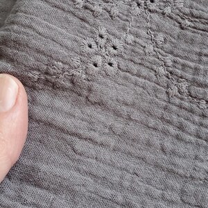 以下が含まれることがあります： 繊細な花柄の刺繍が施された、ダークグレーのしわ加工生地。素材は目に見える織り目があり、柔らかく縮んだような外観で、下端はほつれた縁取りになっており、複雑な刺繍のディテールが際立っています。