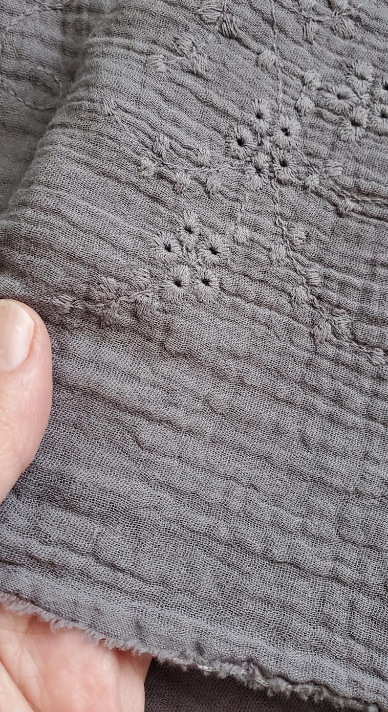 以下が含まれることがあります： 繊細な花柄の刺繍が施された、ダークグレーのしわ加工生地。素材は目に見える織り目があり、柔らかく縮んだような外観で、下端はほつれた縁取りになっており、複雑な刺繍のディテールが際立っています。