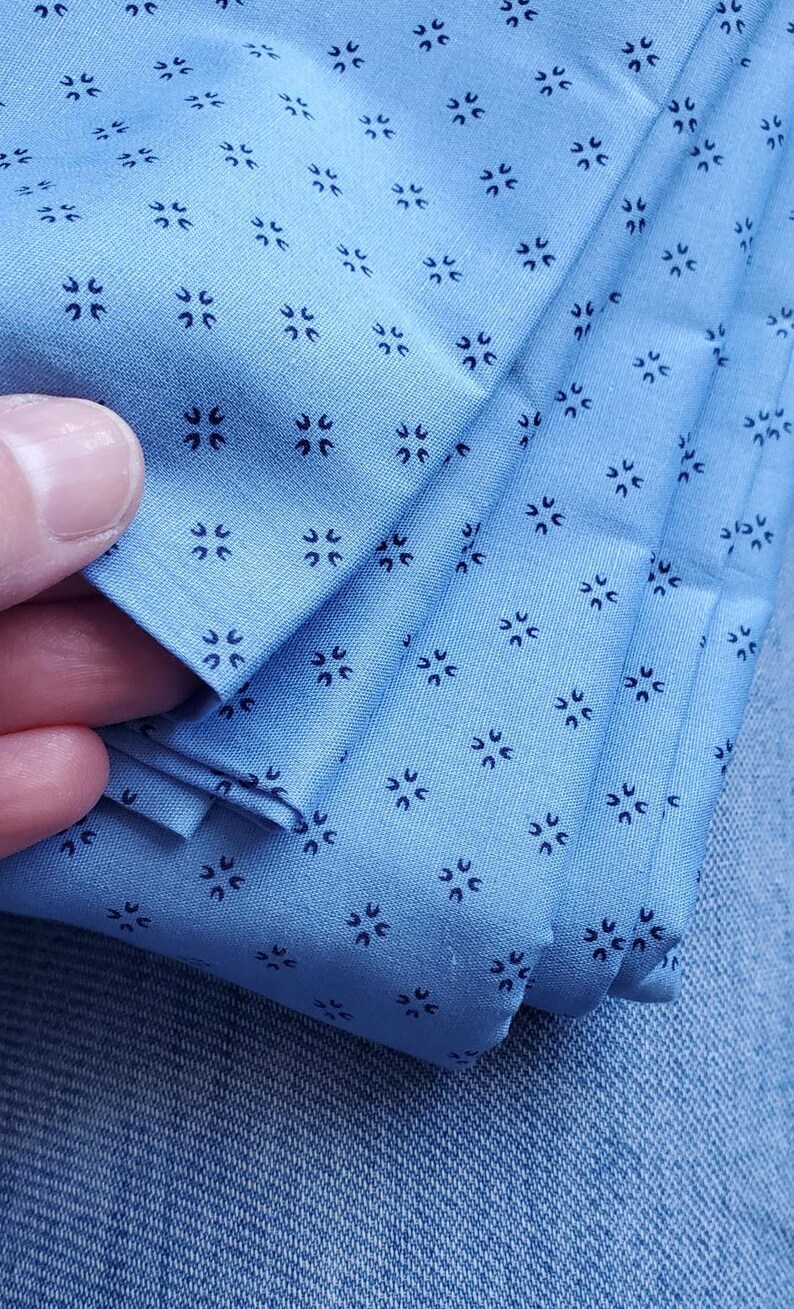 May include: Close-up of folded light blue fabric featuring a repeating pattern of small, dark blue floral designs. The fabric is held, highlighting its texture and colour. The overall theme is textile.