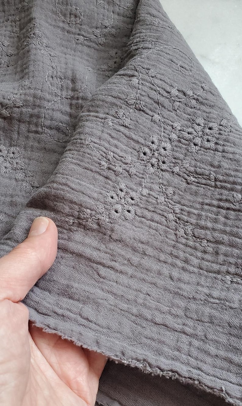 May include: A piece of grey crinkled fabric featuring a delicate tonal embroidered floral pattern. The textile has a soft, textured appearance with a raw, unfinished edge visible along the bottom, showcasing the intricate stitching details.