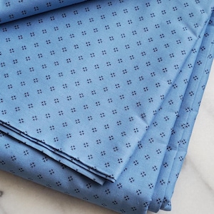 May include: Folded light blue fabric with a repeating pattern of small, dark blue floral shapes. The fabric is neatly stacked, showcasing its texture and design. Suitable for sewing or home decor.