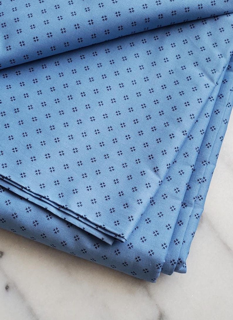 May include: Folded light blue fabric with a repeating pattern of small, dark blue floral shapes. The fabric is neatly stacked, showcasing its texture and design. Suitable for sewing or home decor.