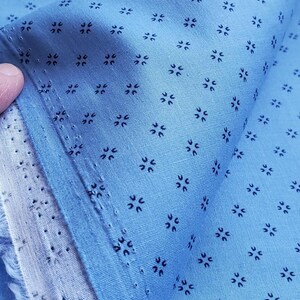May include: Pale blue fabric featuring a repeating pattern of small, dark blue floral motifs. The fabric is folded, revealing its texture and the edge of the material. The fabric is likely cotton or a similar material.