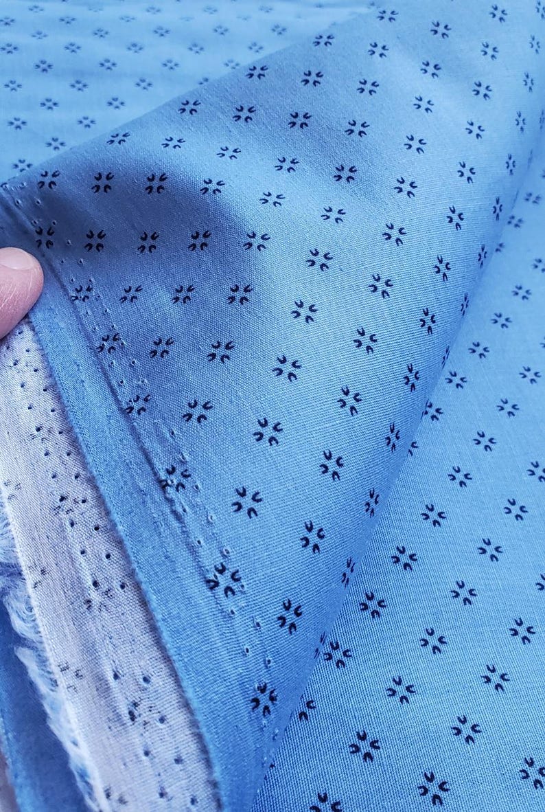 May include: Pale blue fabric featuring a repeating pattern of small, dark blue floral motifs. The fabric is folded, revealing its texture and the edge of the material. The fabric is likely cotton or a similar material.