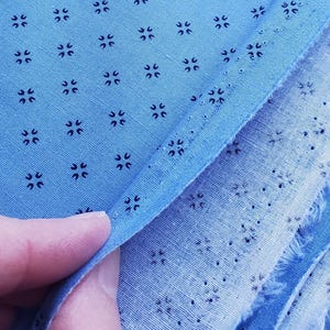 May include: Close-up of light blue fabric with a repeating pattern of small, dark blue floral-like shapes. The fabric is folded, revealing a raw edge and the texture of the material. The image highlights the detailed pattern and colour of the textile.