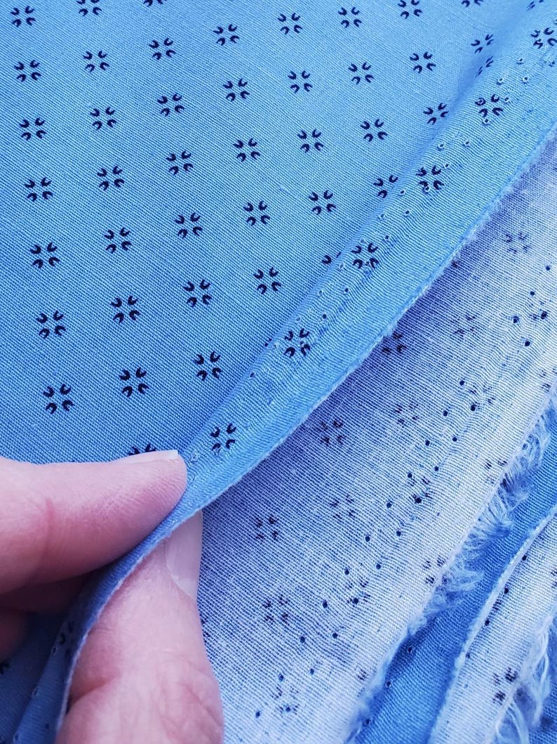 May include: Close-up of light blue fabric with a repeating pattern of small, dark blue floral-like shapes. The fabric is folded, revealing a raw edge and the texture of the material. The image highlights the detailed pattern and colour of the textile.