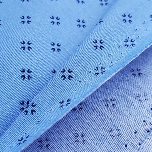 May include: Close-up of light blue fabric with a repeating pattern of small, dark blue floral-like designs. The fabric is folded, revealing the texture and the intricate details of the pattern. The fabric has small holes.