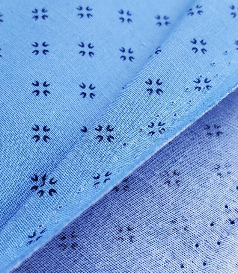 May include: Close-up of light blue fabric with a repeating pattern of small, dark blue floral-like designs. The fabric is folded, revealing the texture and the intricate details of the pattern. The fabric has small holes.