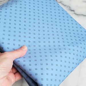 May include: A folded piece of light blue fabric with a repeating pattern of small, dark blue floral-like shapes. The fabric is held in a hand, showcasing its texture and colour. The fabric appears to be cotton or a similar material, suitable for sewing projects.