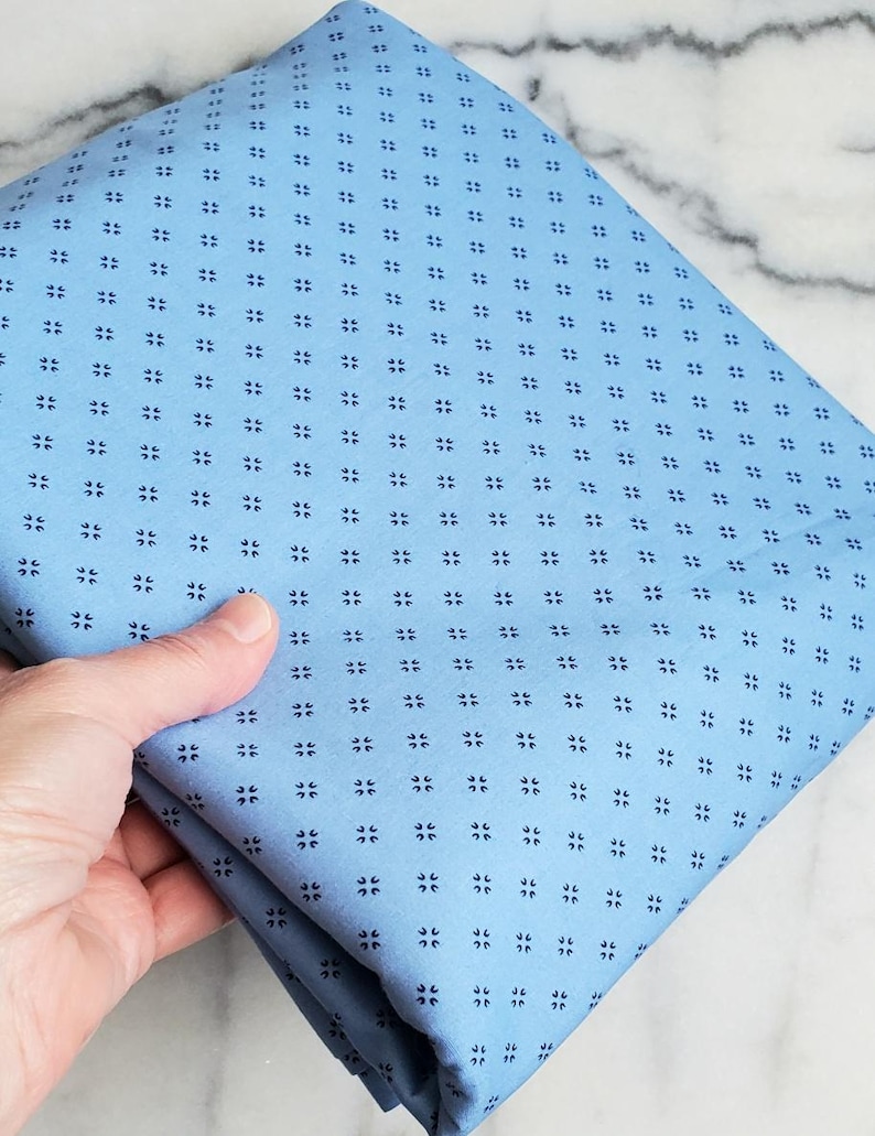 May include: A folded piece of light blue fabric with a repeating pattern of small, dark blue floral-like shapes. The fabric is held in a hand, showcasing its texture and colour. The fabric appears to be cotton or a similar material, suitable for sewing projects.