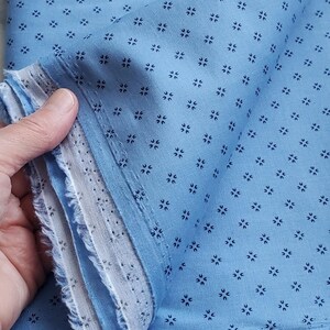 May include: A close-up of blue fabric with a repeating pattern of small, dark blue floral shapes. The fabric has a light blue background and a frayed edge. The fabric is folded. The image shows the edge of the fabric.